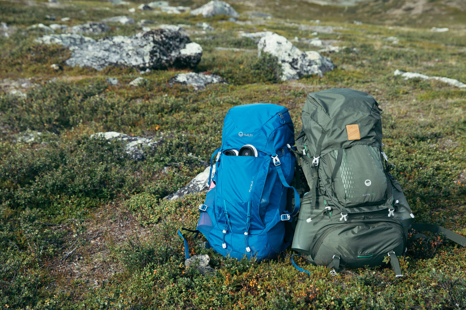 Two Halti Inari 75 Backpacks, one blue and one green, sit on grassy terrain with rocks in the background.
