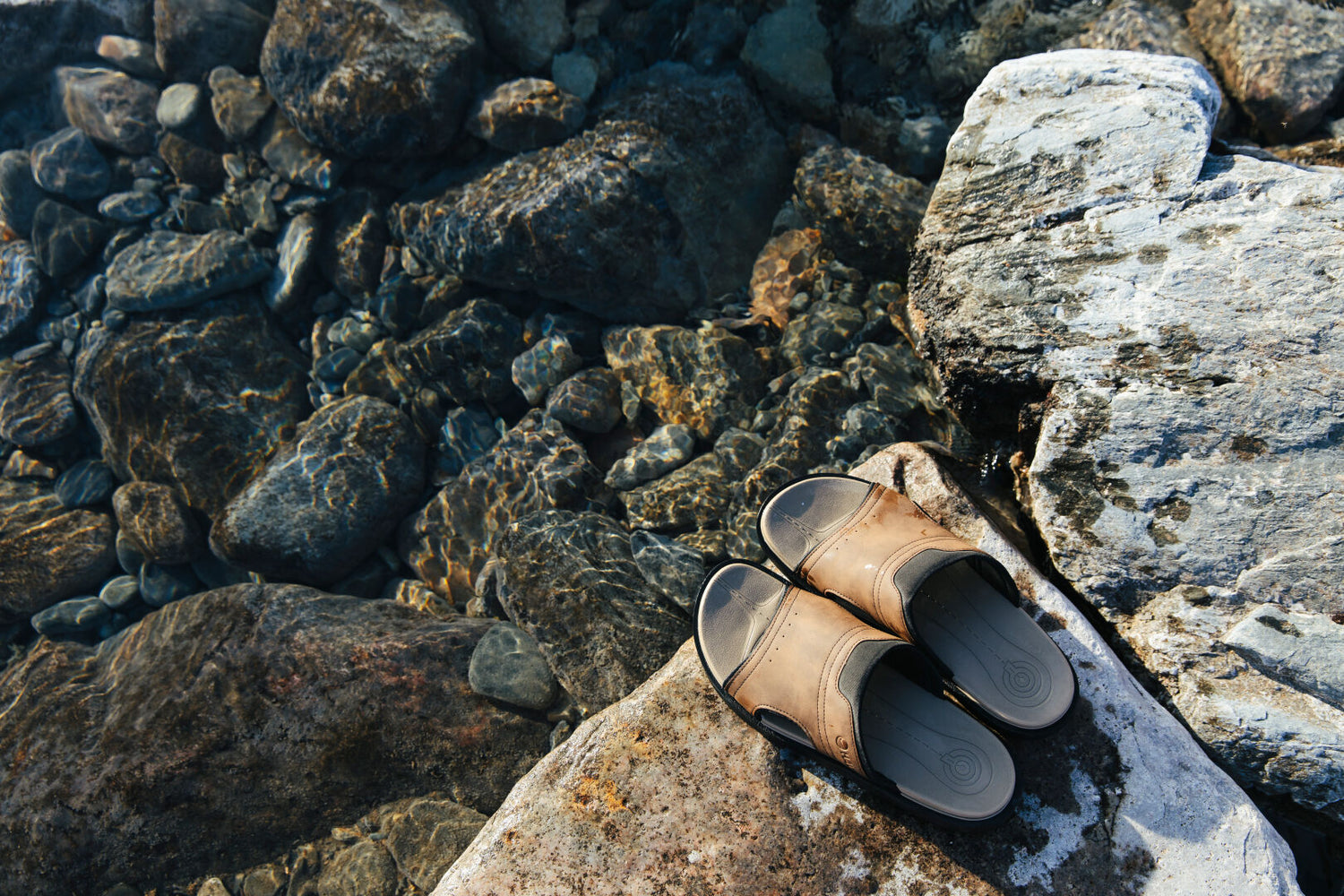 Halti Jorah Mens Sandals, made of synthetic leather with a lightweight Phylon sole, rest on a rock by clear water.