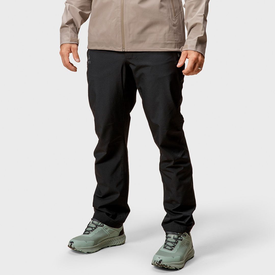 A person in a beige jacket and Halti Vuoksi DX Shell Pants stands against a plain background, wearing light green hiking shoes.
