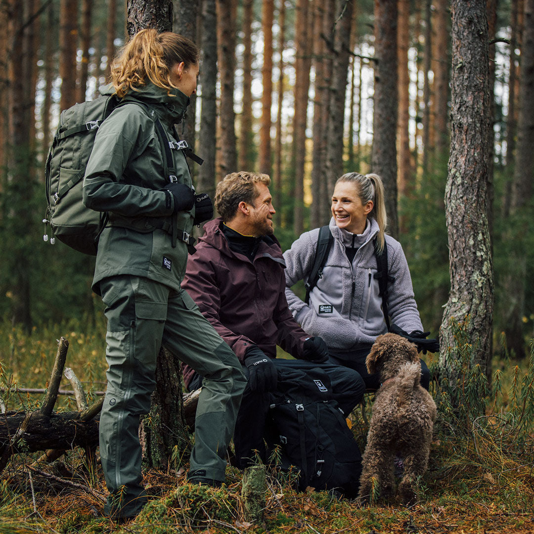 Three women in Halti Hiker 3L DX Ventilated Pants chat in a pine forest with a small brown dog nearby.
