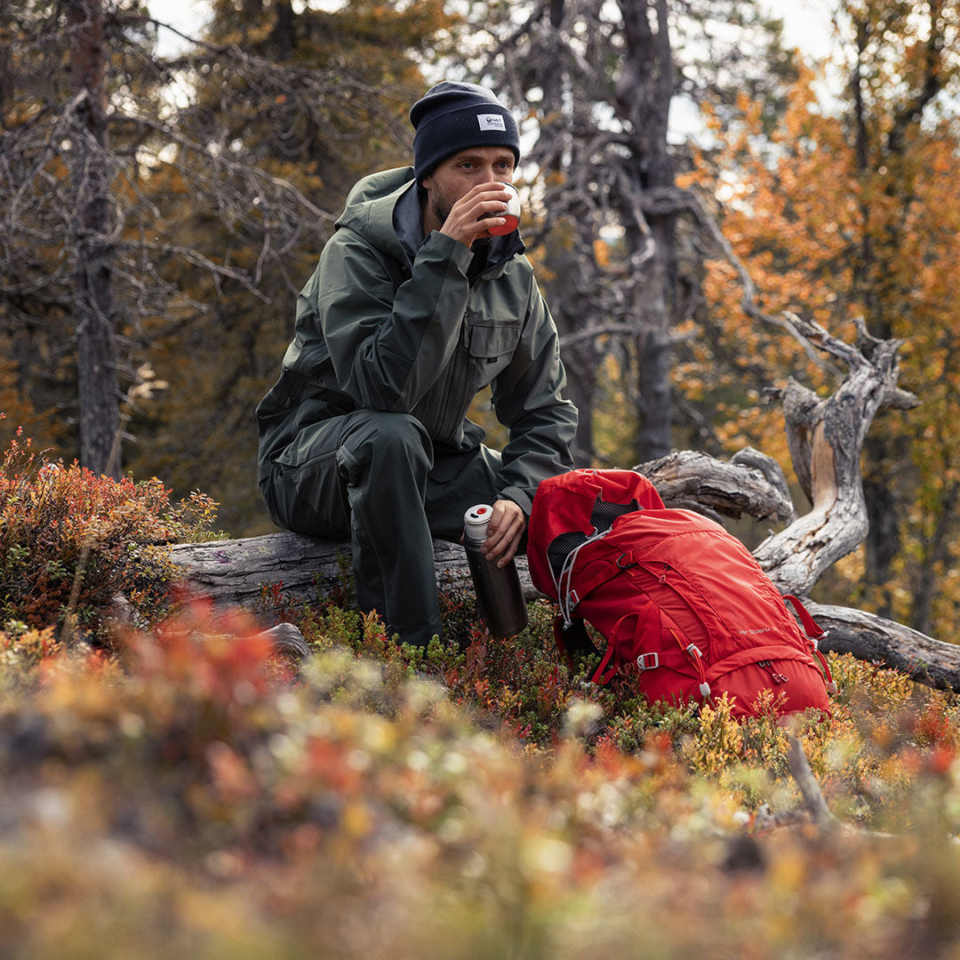 Man in Halti Hiker 3L DX Ventilated Pants sits on a log with a cup; red backpack and trees in the background.