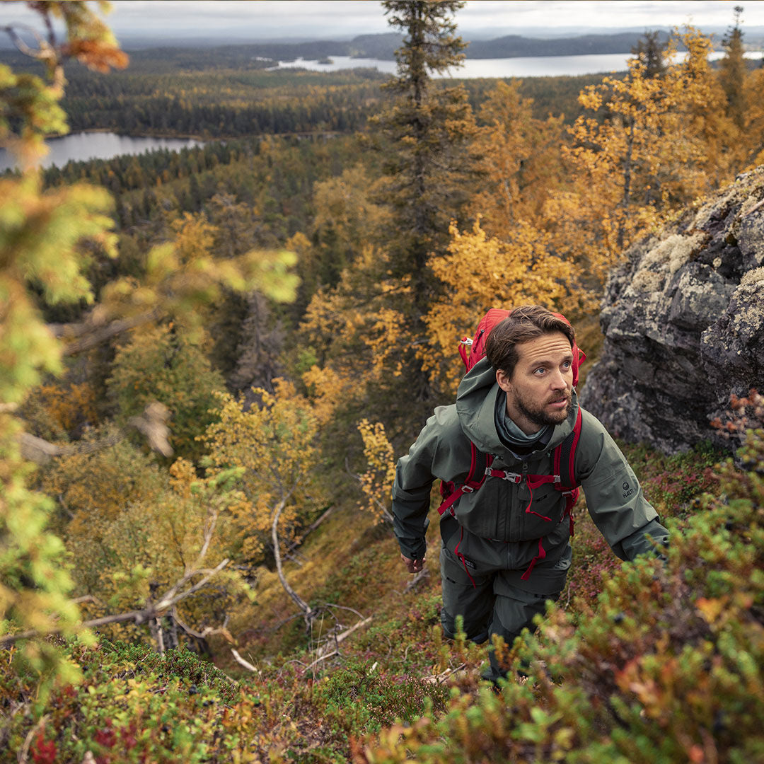Man in a Halti Hiker 3L DX Ventilated Jacket and red backpack hikes up a rocky hill in an autumn lakeside forest.