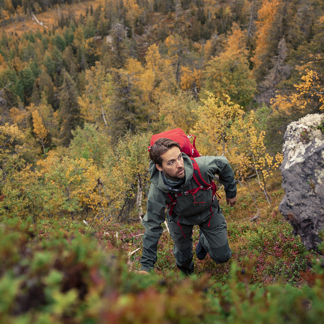 A man wears the Halti Hiker 3L DX Ventilated Jacket climbing a grassy, steep hill with autumn trees in the background.