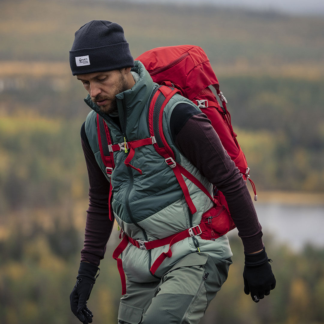 A man in Halti Hiker 3L DX Ventilated Pants and a red backpack hikes a scenic trail with trees and water behind him.
