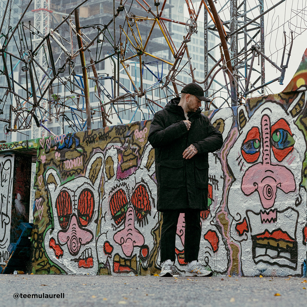 A person in a Halti Bergga Mens DrymaxX Winter Jacket stands before a graffiti wall with cartoon faces and metal art above.