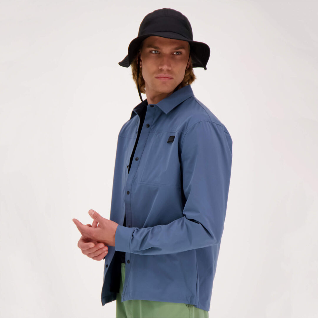 A person in Haltis Bluusi Unisex Shirt, black hat, and light green pants stands against a plain, light background.