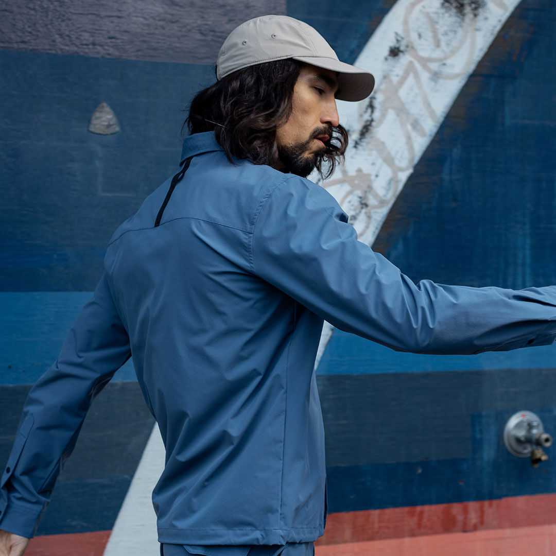Man wears Halti Bluusi Unisex Shirt and beige cap, standing sideways by a blue wall with white and red accents.