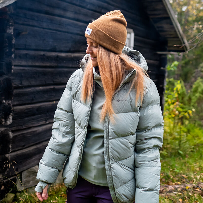 Woman in Halti Versant Down Jacket stands outside by a dark wooden cabin and green foliage, wearing a brown beanie.