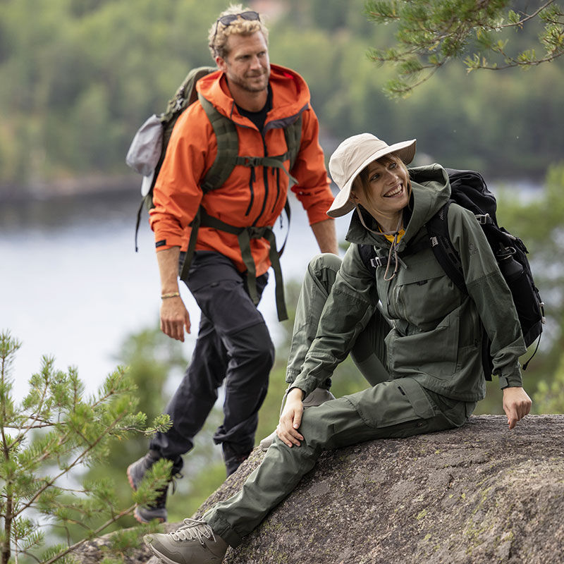 Two women in Halti Hiker Womens Ventilated Pants explore lakeside, one seated and one standing among the trees.