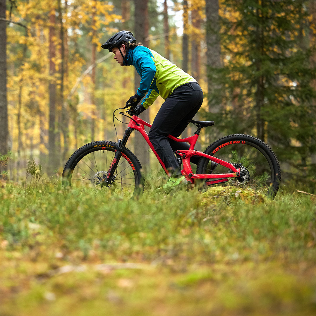 Wearing Halti Suunta Mens Hybrid Pants, a cyclist rides a red mountain bike through a green, wooded forest.