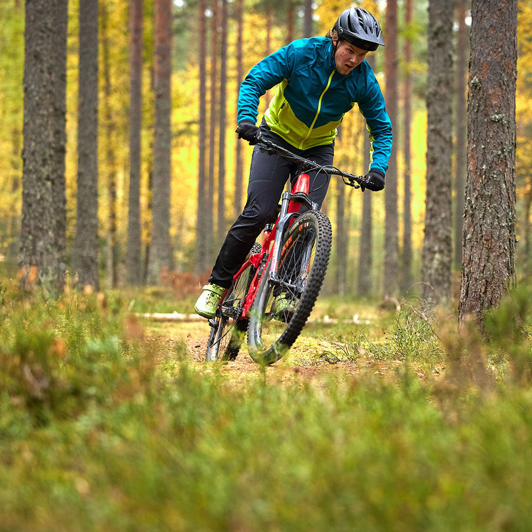 A man in Halti Suunta Mens Hybrid Pants rides a mountain bike on a forest trail surrounded by lush greenery.