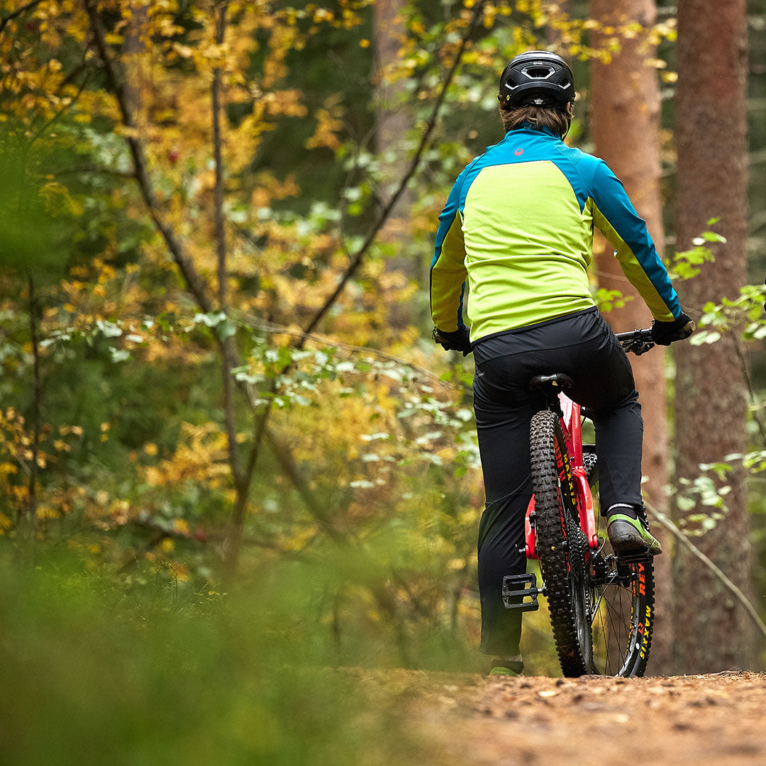 A person in Halti Suunta Mens Hybrid Pants and a helmet rides a mountain bike on a dirt trail through an autumn forest.