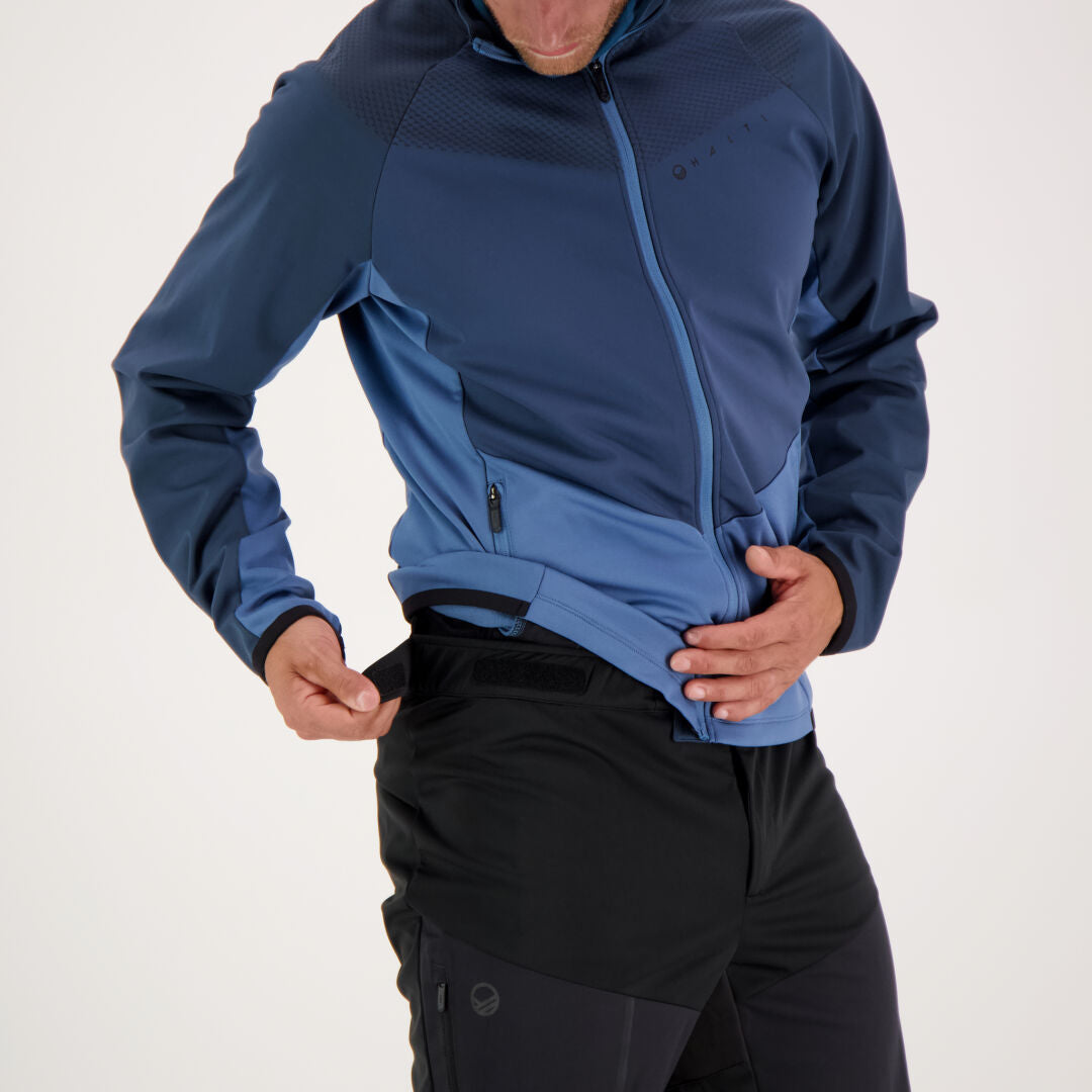 A man in a blue jacket adjusts the waistband of Halti Suunta Mens Hybrid Pants against a plain light background.