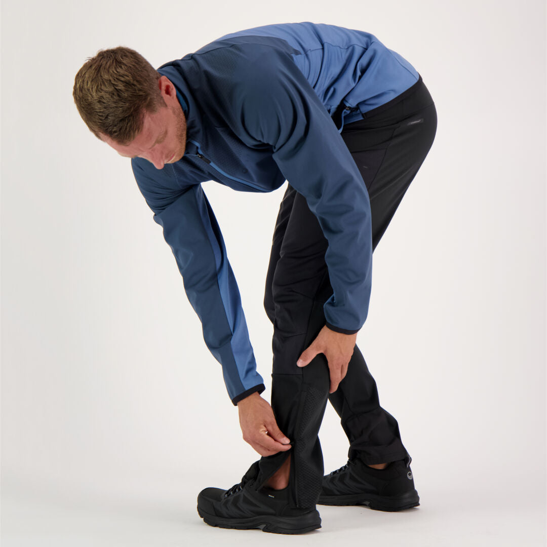 A person in a blue jacket unzips the lower leg of Halti Suunta Mens Hybrid Pants over black shoes.