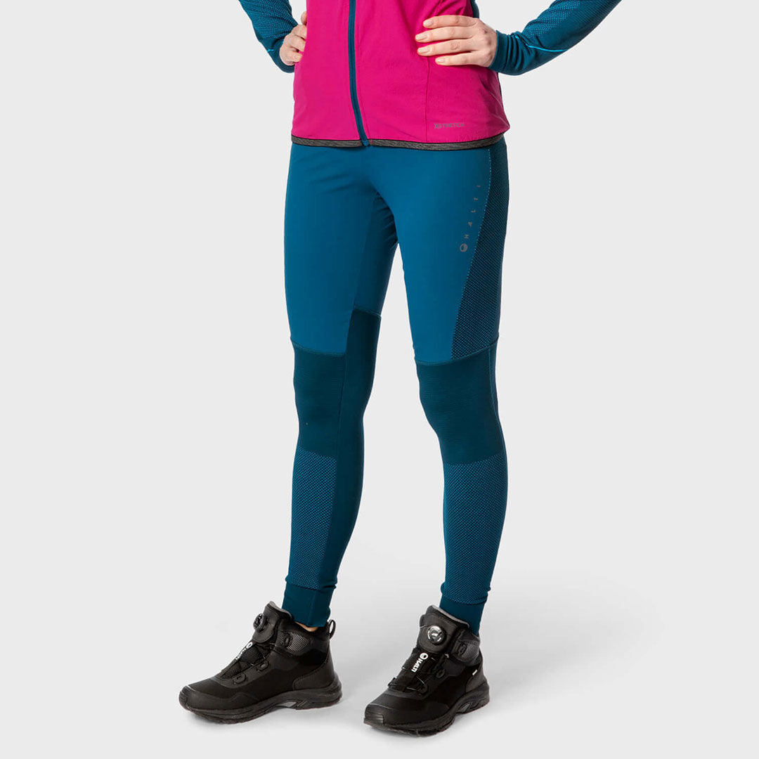 A woman in Halti Tracks Seamless Tights stands in black boots and a pink-blue jacket against a light background.