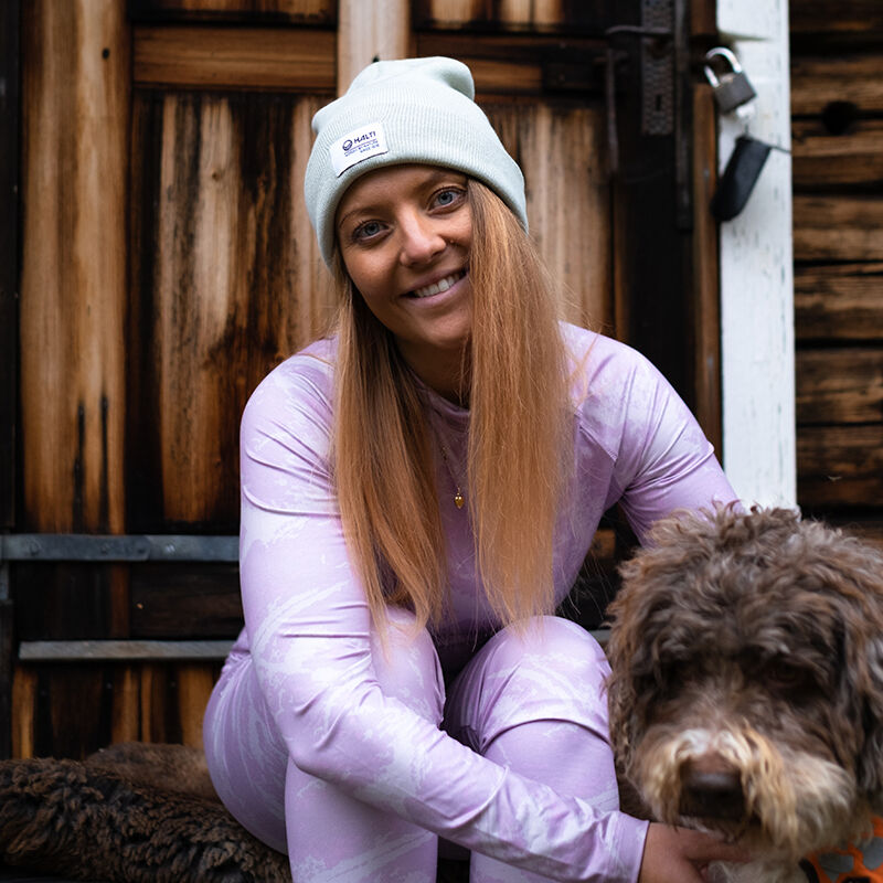 A woman in the Halti Windfire Baselayer set and gray beanie sits on steps, petting a brown curly-haired dog.