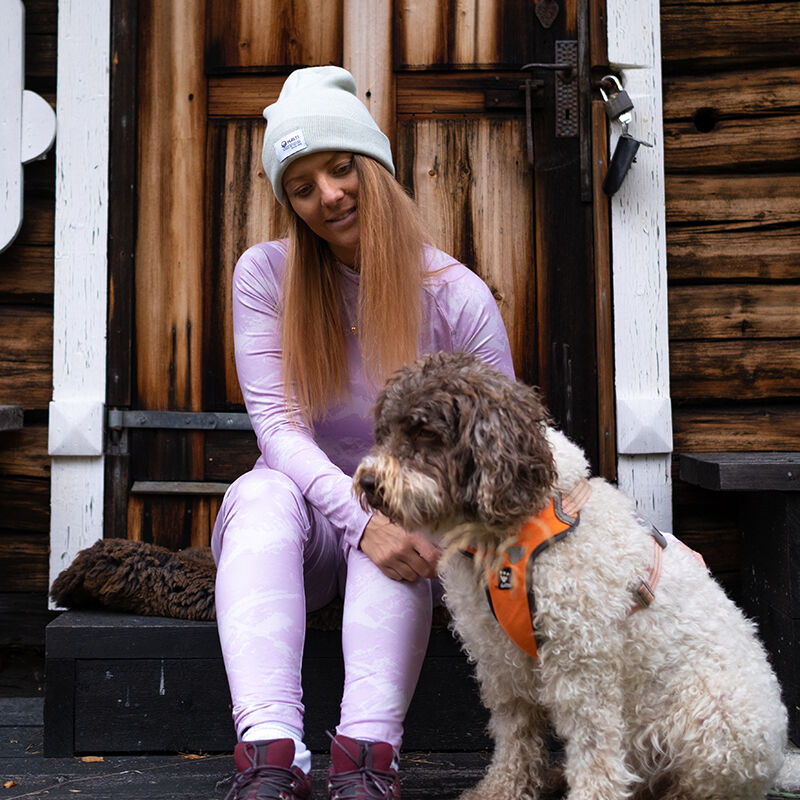 A woman in a Halti Windfire Baselayer set sits on wooden steps beside a curly-haired dog in an orange harness.