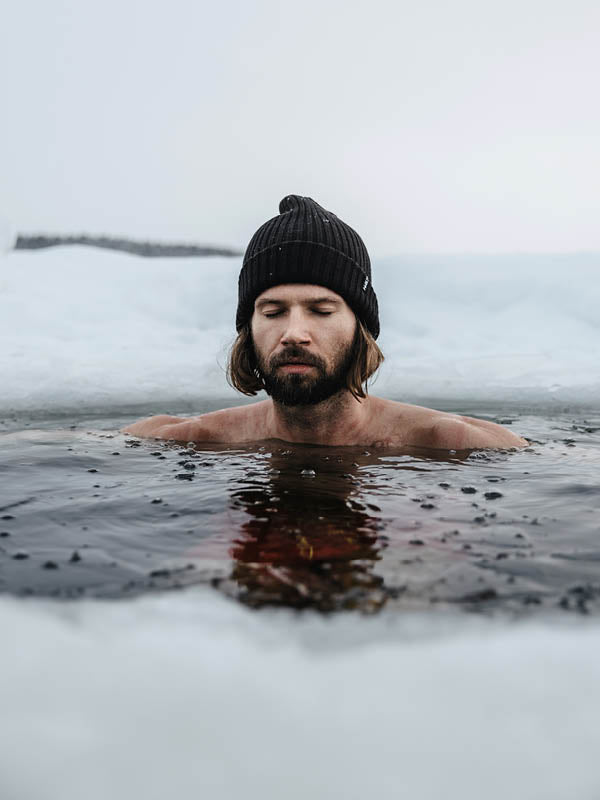 A man with shoulder-length hair and a black beanie is submerged up to his chest in icy water, surrounded by snow and ice, with his eyes closed and a calm expression.