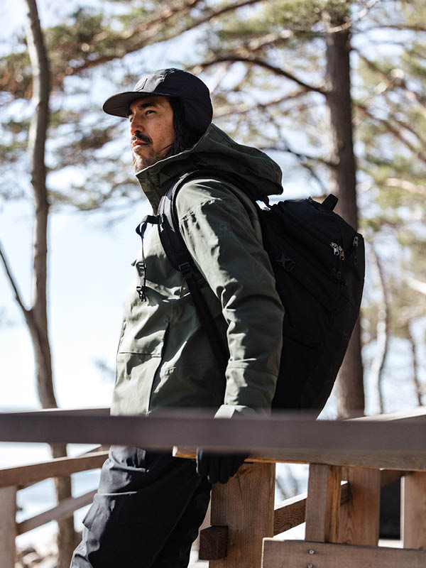 A person wearing a dark green jacket, black cap, and black backpack stands outdoors on a wooden structure, with trees and sunlight in the background.
