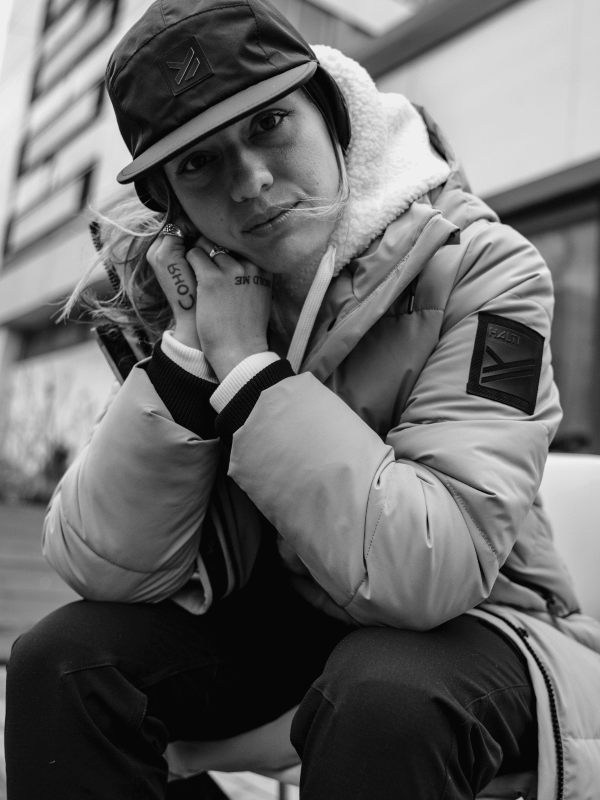 A person wearing a puffer jacket, hoodie, and cap sits outdoors, leaning forward with one hand on the side of their face, looking at the camera. The image is in black and white.