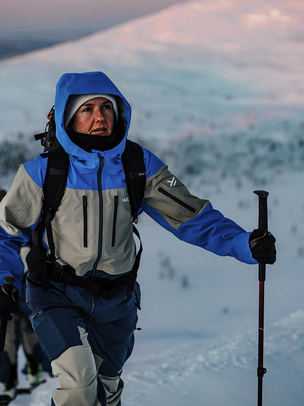 A person wearing a blue and gray winter jacket, gloves, and a hood hikes uphill in a snowy mountain landscape, holding a trekking pole. The background shows snow-covered slopes.