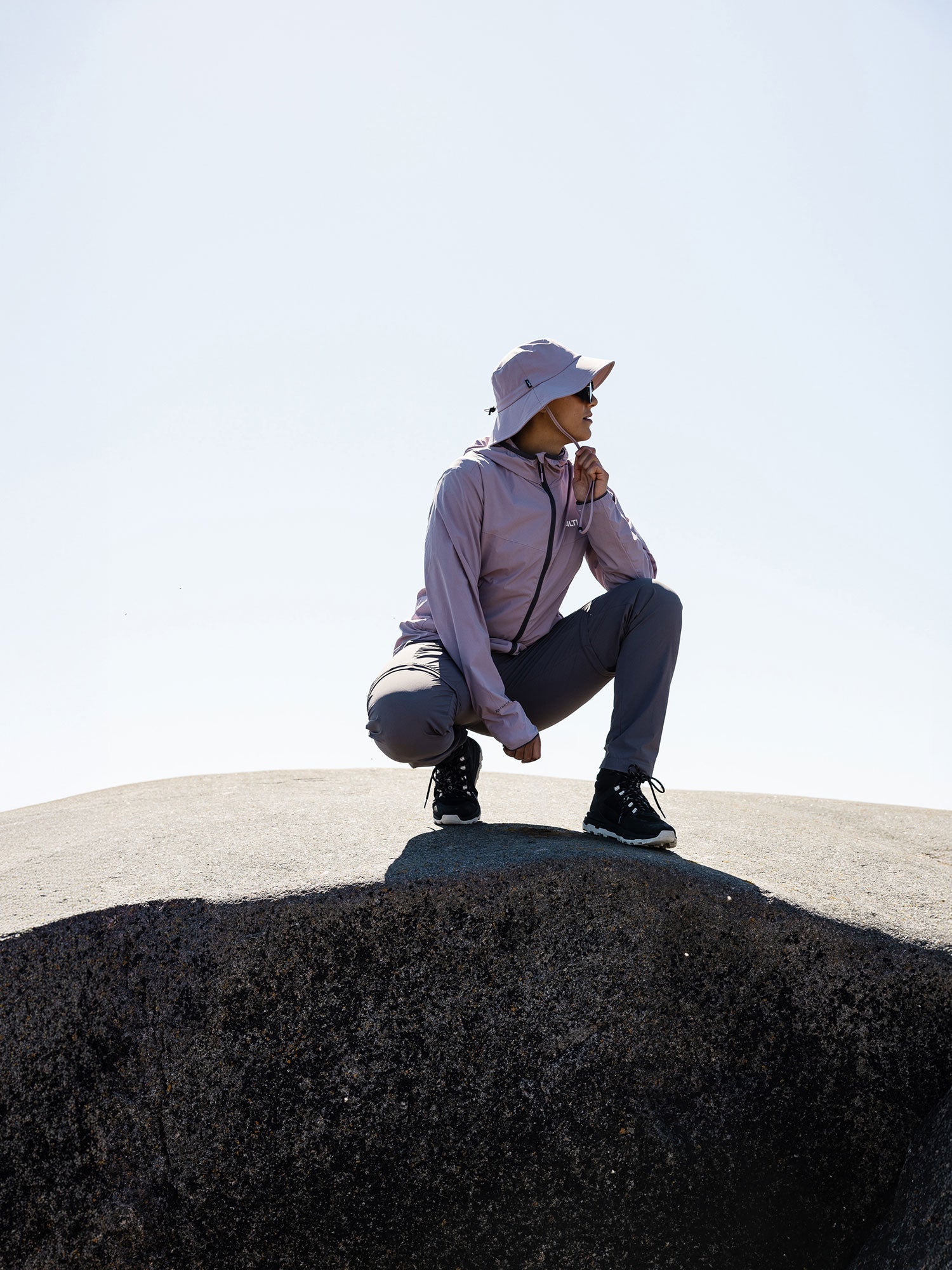 A person in a light purple jacket and hood sits on a rock outdoors. Text reads HALTI, Cool on skin, blocks the sun, and UPF 90+ ultimate sun protection. Sunglasses reflect a landscape.