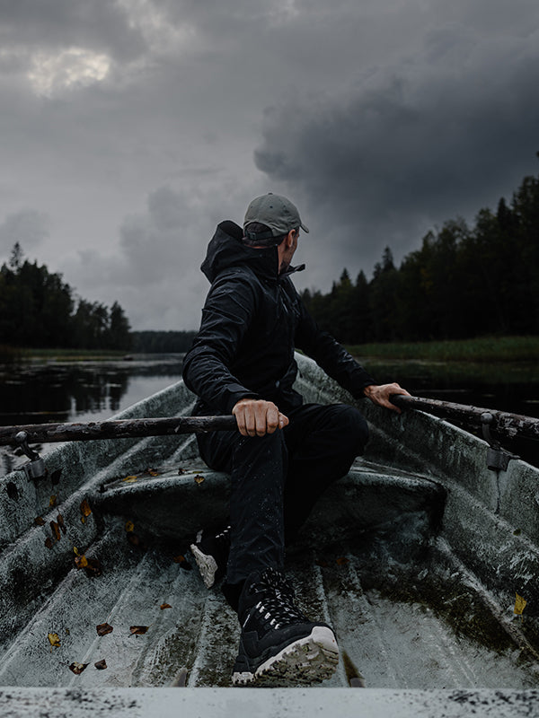 A person wearing dark outdoor clothing and a cap rows a small, weathered boat on a calm river, surrounded by trees under a cloudy, overcast sky.