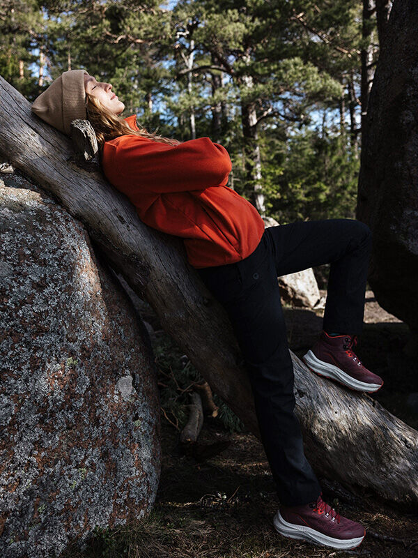 A woman in an orange jacket leans on a tree, wearing Halti Rapid DX Mid Outdoor Shoes in a rocky, wooded area.