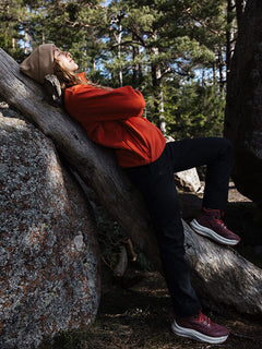 A woman in an orange jacket leans on a tree, wearing Halti Rapid DX Mid Outdoor Shoes in a rocky, wooded area.