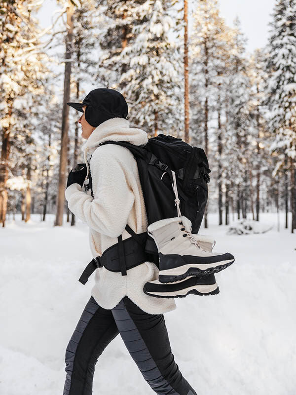 A hiker in winter gear carries a backpack with Halti Tornio 2 DX Winterboots attached in a snowy forest.
