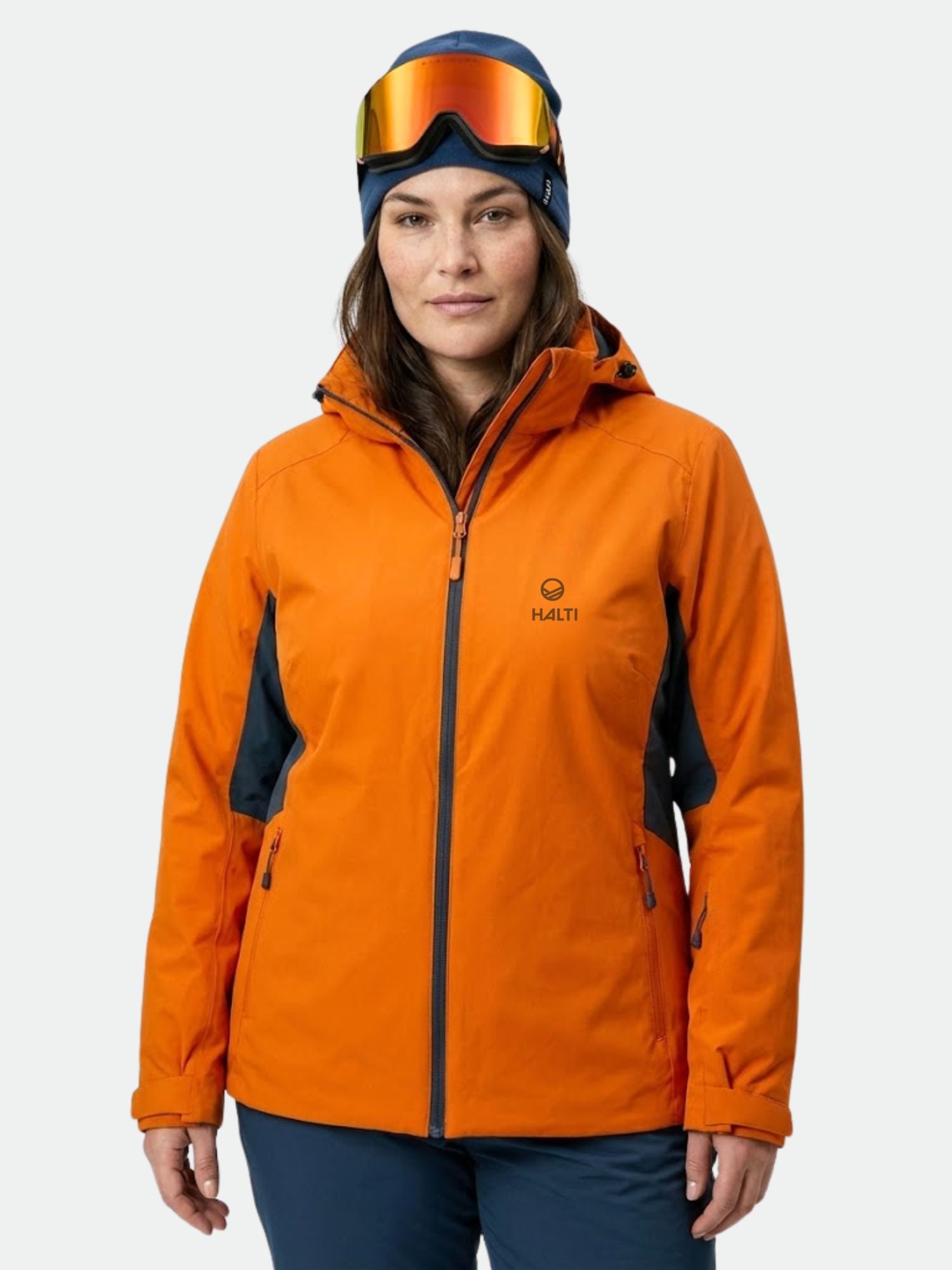 A woman wears the Halti DX Ski Jacket Women's Plus in orange with ski goggles, facing forward against a white background.