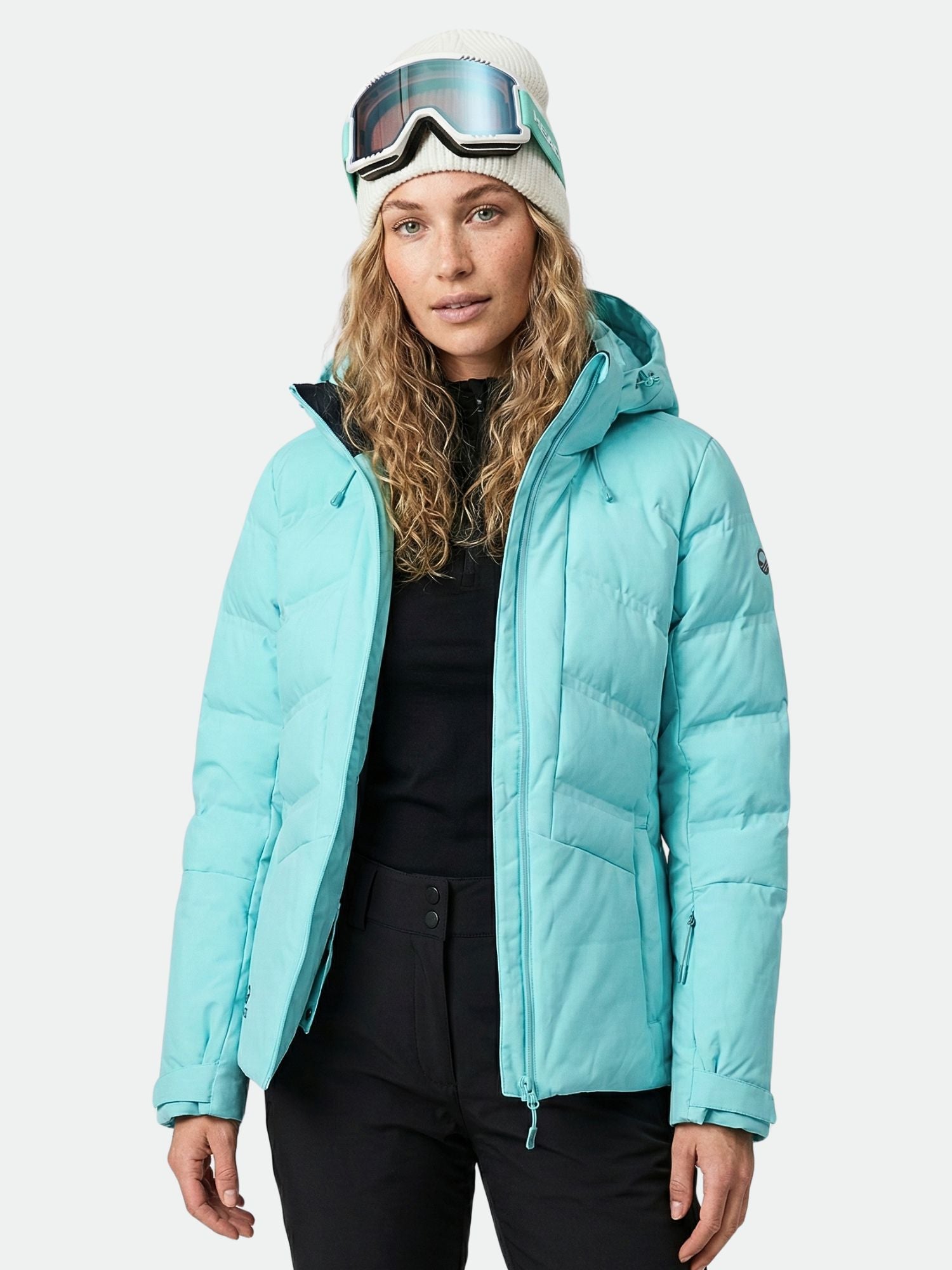 A woman wears the Halti Nordic Arcty Ski Jacket in light blue, black pants, white beanie, and ski goggles facing forward.