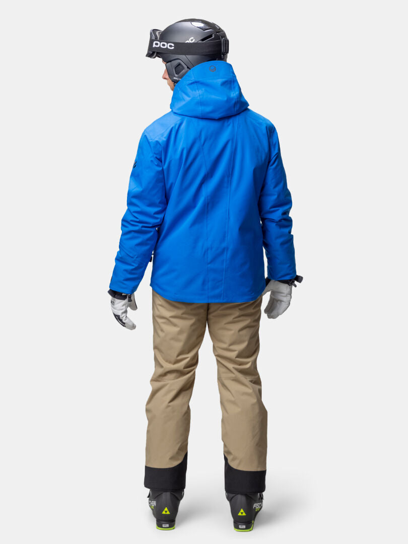 Man in a Halti Vertica DX Ski Jacket (blue), tan pants, ski boots, gloves, and helmet, standing facing away.