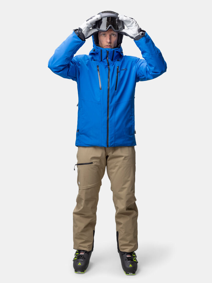 Wearing a breathable Halti Vertica DX Ski Jacket in blue, beige pants, and boots, he adjusts ski goggles on his head.