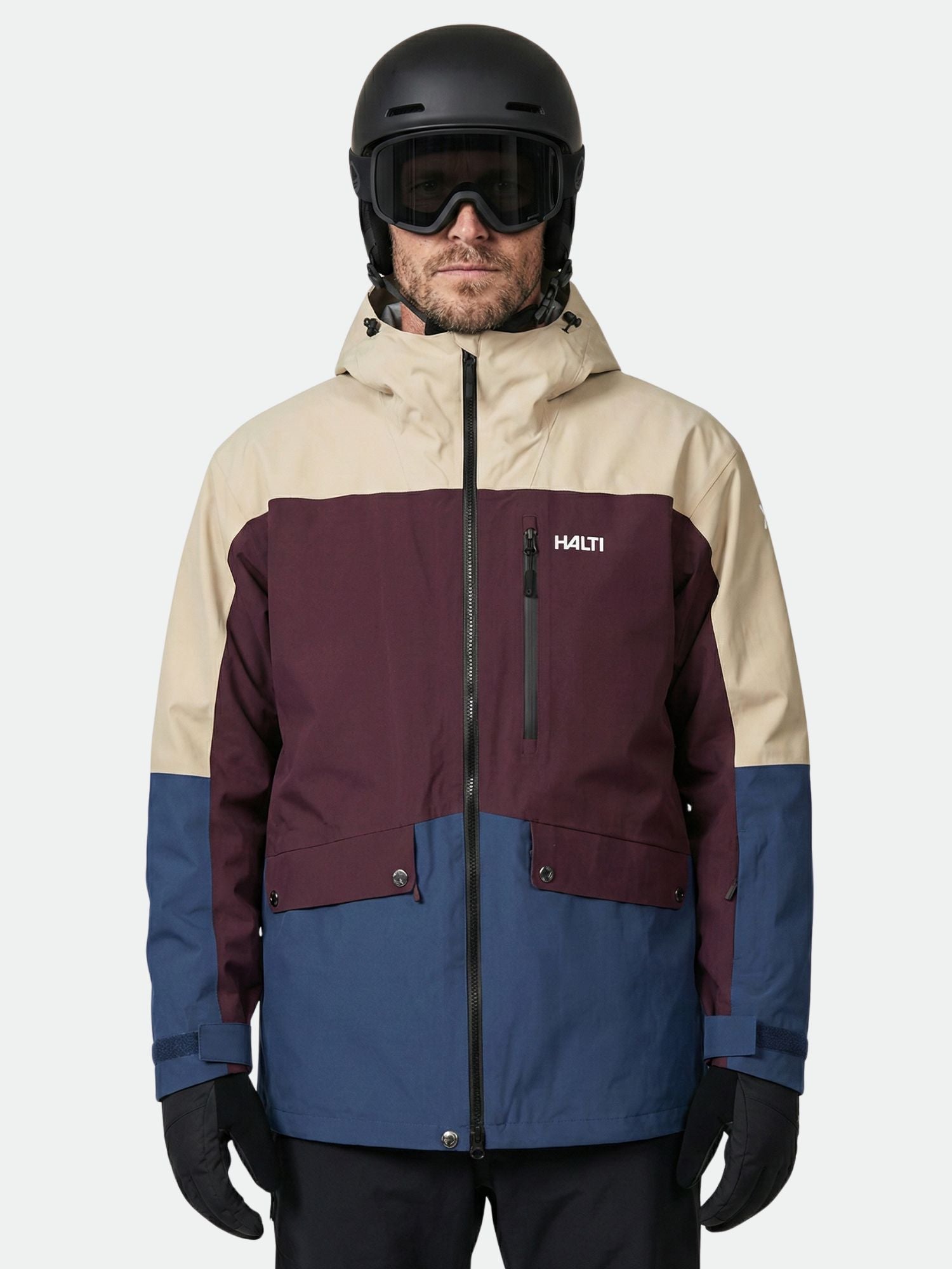 A person in a Halti Settler 3L DX Ski Jacket (men's) in beige, burgundy, and blue with black pants, gloves, helmet, and goggles.