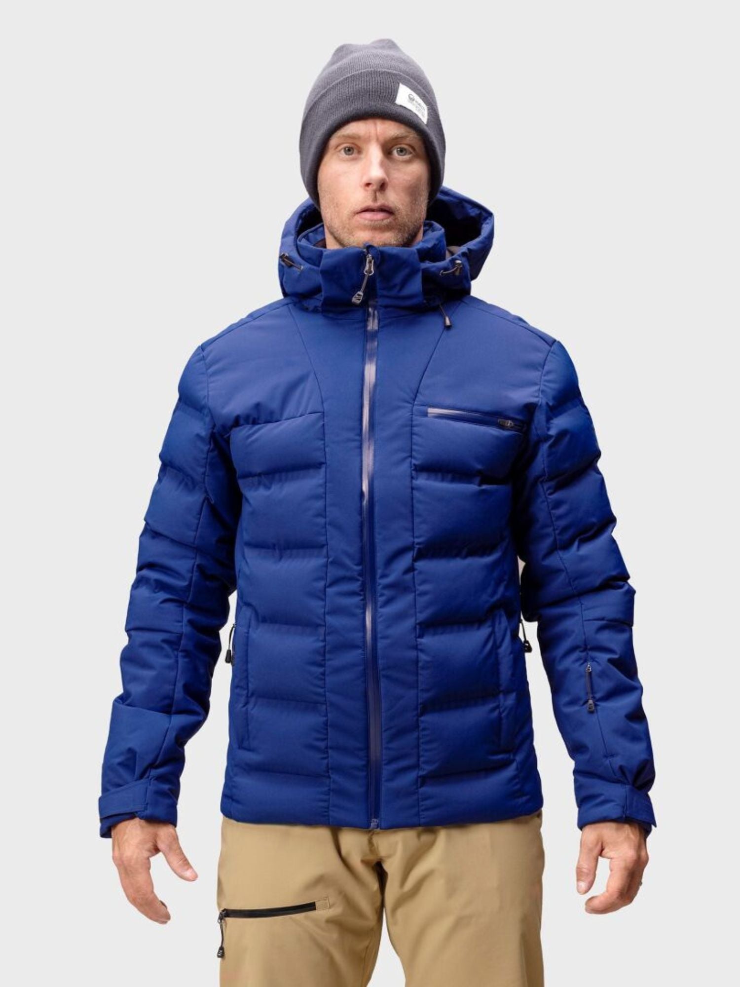 A person wears a Halti Nordic Arcty Ski Jacket, tan pants, and a gray beanie against a plain light background.