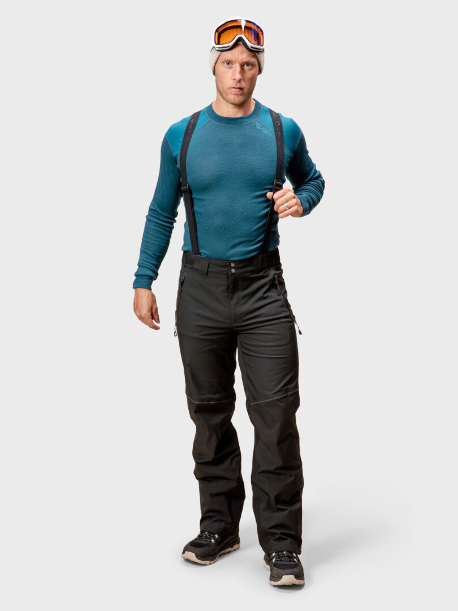A man wears Halti Vertica DrymaxX Ski Pants, a teal long-sleeve shirt, beanie, and goggles, facing forward.