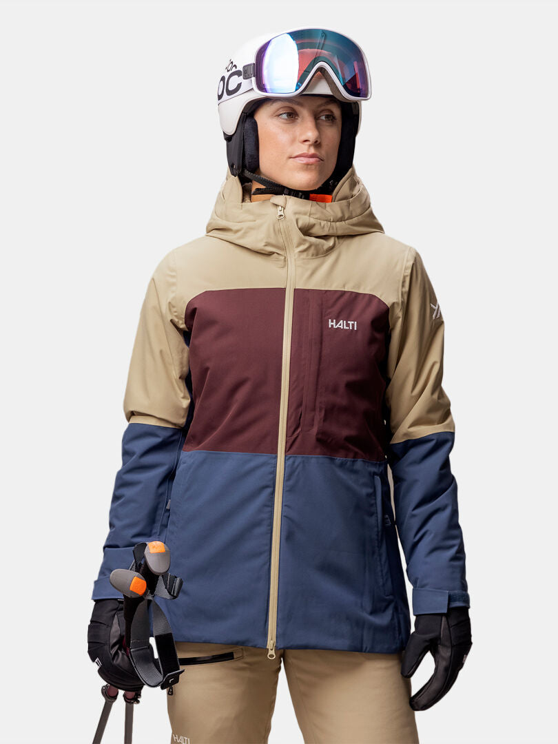 A woman wears the Halti Gracer DX Ski Jacket, helmet, goggles, and gloves, holding ski poles against a plain background.