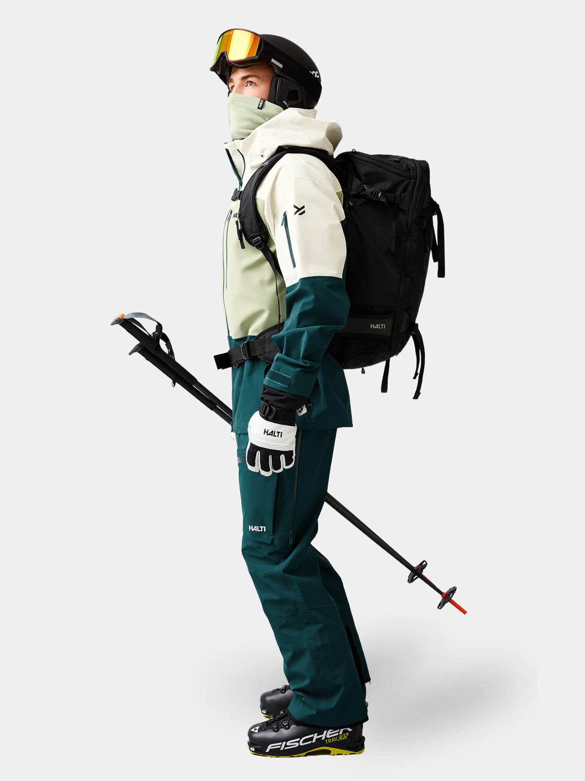 A skier wears a cream and teal Halti Rizle Unisex 3L DX Ski Jacket, helmet, goggles, and a backpack with ski poles.