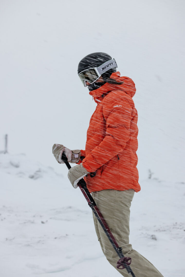A woman in an orange jacket wears Halti Carvey II DX Ski Pants, helmet, and holds ski poles amid snowy winds.