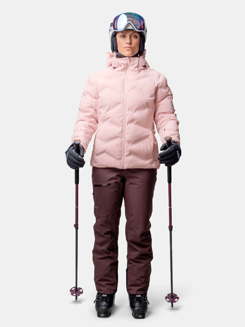 A person in a Halti Nordic Arcty II Ski Jacket for women, brown pants, helmet, and goggles holds ski poles against a plain background.