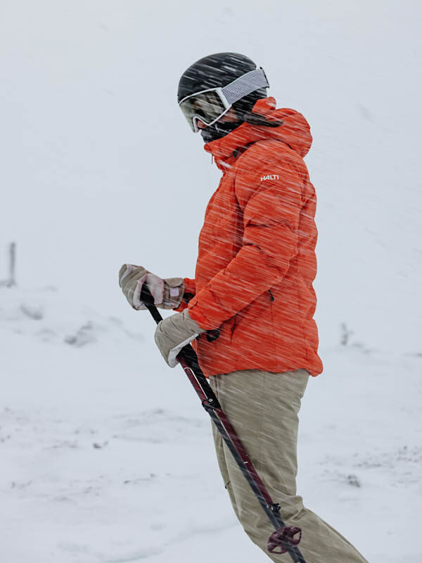 Wearing the Halti Nordic Arcty II Ski Jacket, a woman stands in snow and wind with ski poles and goggles.