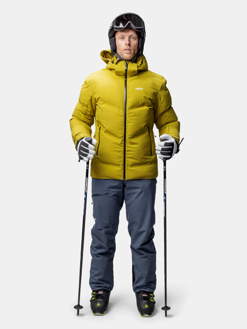 A person in a yellow Halti Nordic Arcty II Ski Jacket, blue pants, helmet, goggles, and gloves stands with ski poles.