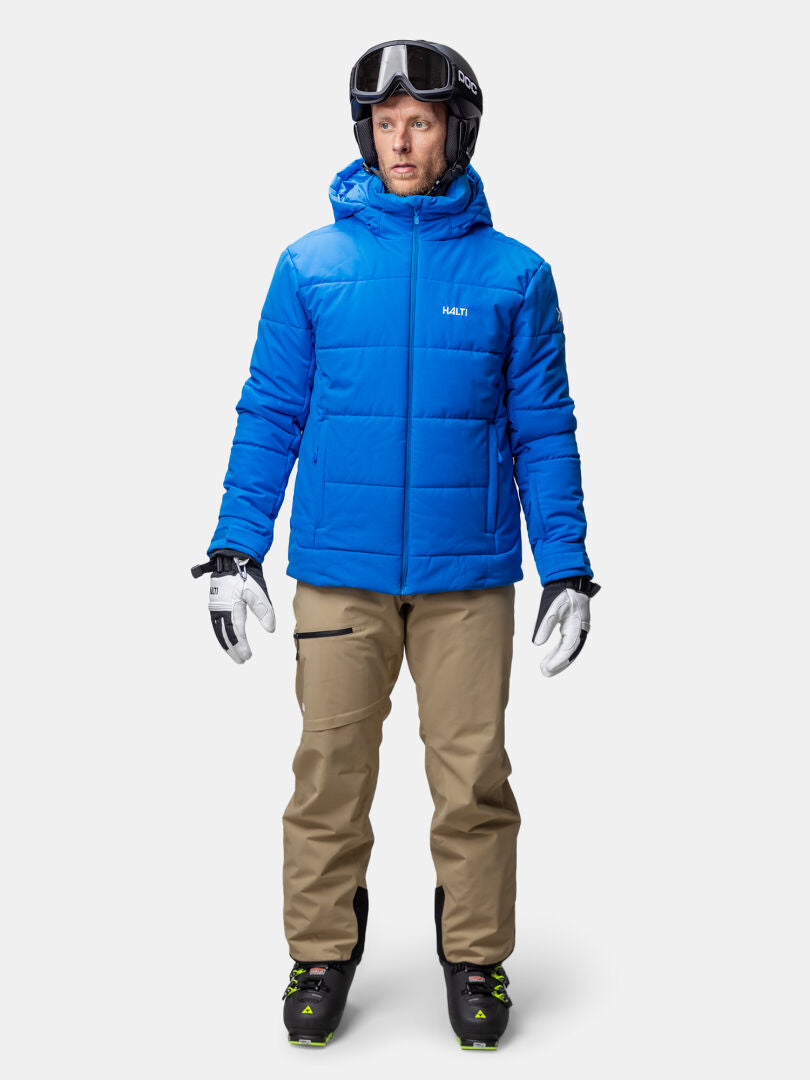 A man in a Halti Ascale Puffer Ski Jacket, beige ski pants, white gloves, boots, helmet, and goggles faces forward.