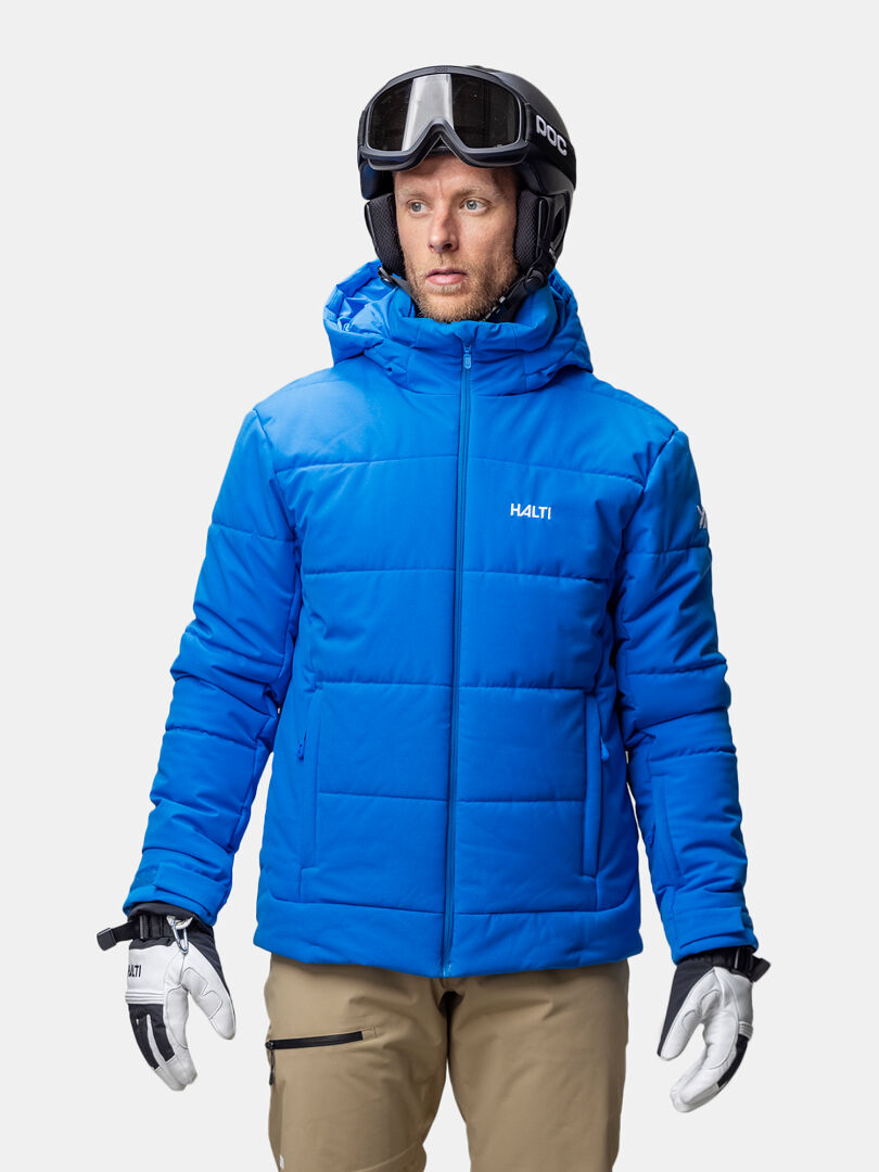 A man wears a Halti Ascale Puffer Ski Jacket, tan pants, gloves, and a helmet with goggles against a plain background.