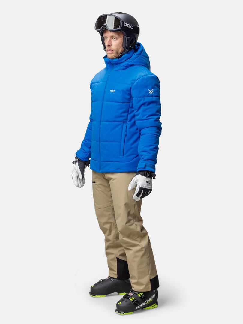 A man wears the Halti Ascale Puffer Ski Jacket, beige pants, ski boots, gloves, and a black helmet with goggles.