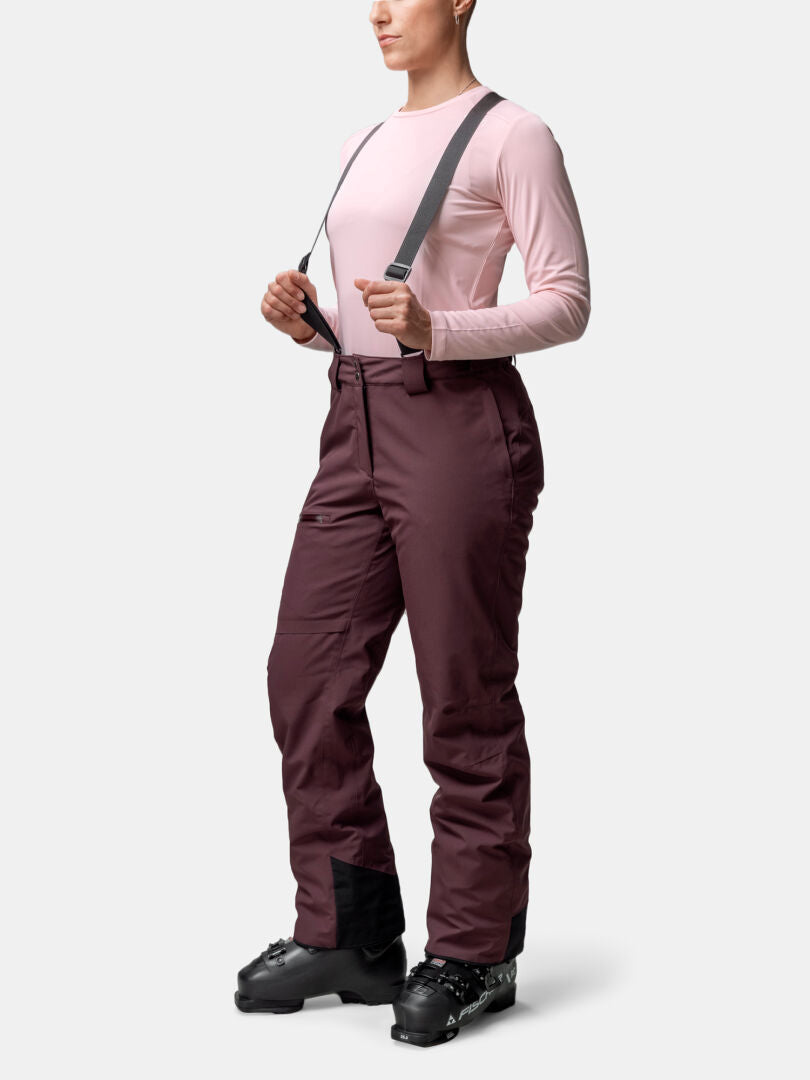 A person wears pink long sleeves, Halti Carvey II DX women’s ski pants with suspenders, and black ski boots forward.