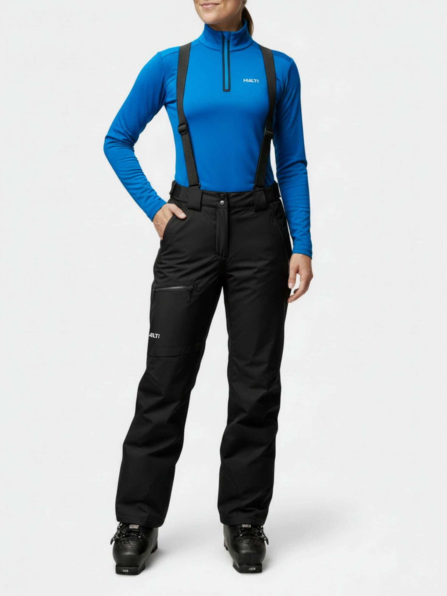 Woman in Halti Carvey II DX Ski Pants and ski boots, blue long-sleeve top, black suspenders; white background.
