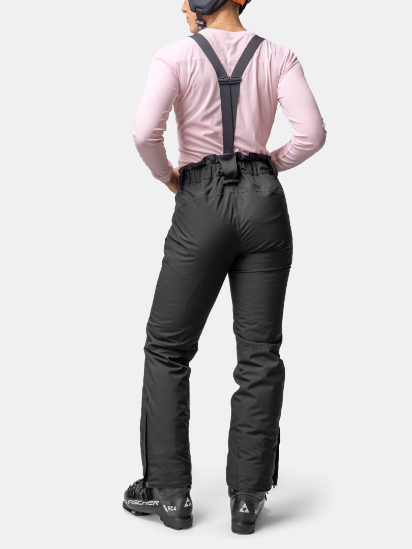 Person wearing a pink long-sleeve shirt, Halti Glades II DX Women's ski pants with suspenders, ski boots, and helmet.