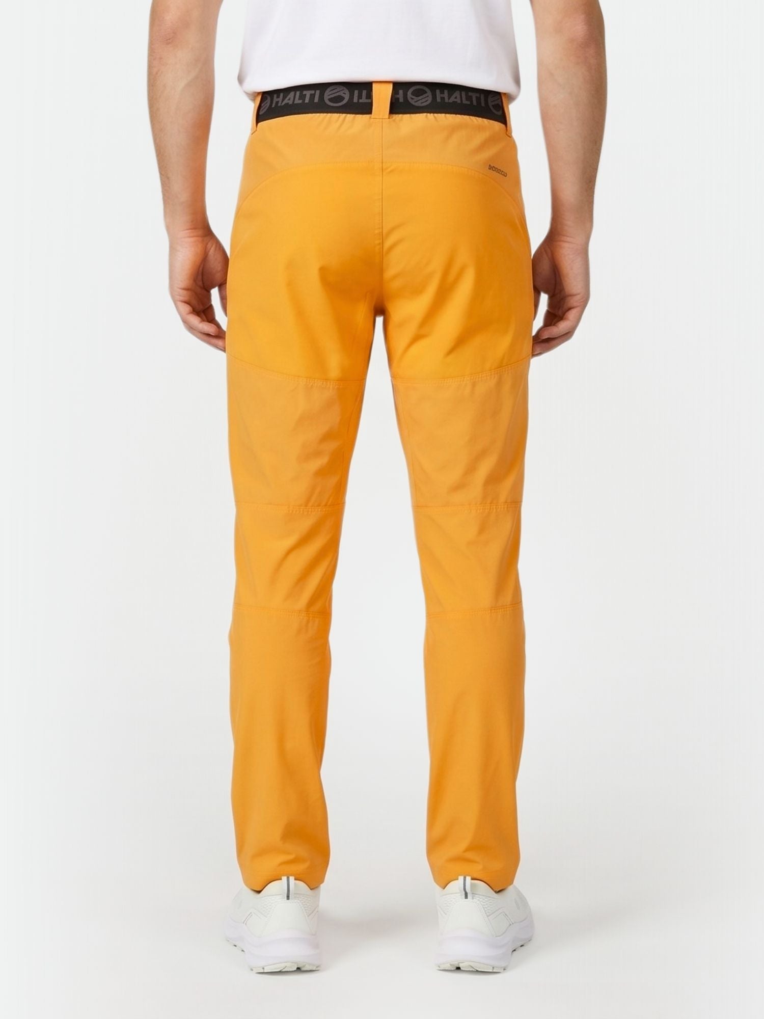 A person in Halti Pallas Men's X-stretch Lite Pants (orange), a white tee, and sneakers, seen from the back on white.
